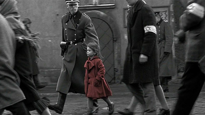 A girl in a red coat (Oliwia Dąbrowska) walks with a concentrated expression, hands in her pockets, surrounded by Jewish men in coats and armbands marked with Stars of David, as an SS officer glares at them while walking in Schindler's List