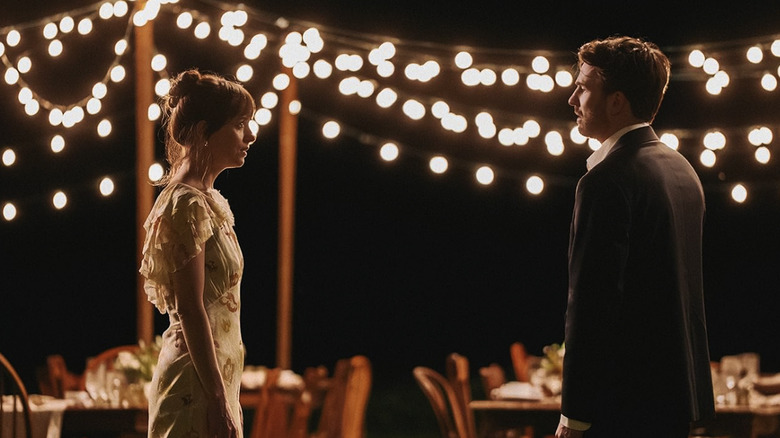 Lucy and John stand in profile with string lights behind them in Materialists