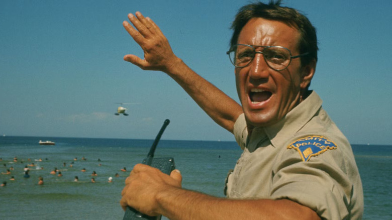 Chief Brody (Roy Scheider) carries a walkie-talkie in one hand and raises his other arm as he shouts, with the beach and a helicopter in the background in Jaws