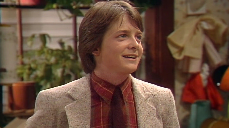 A close-up of Alex P. Keaton (Michael J. Fox) with a bright smile and wearing a beige blazer and red plaid shirt, in front of a cluttered home background in Family Ties