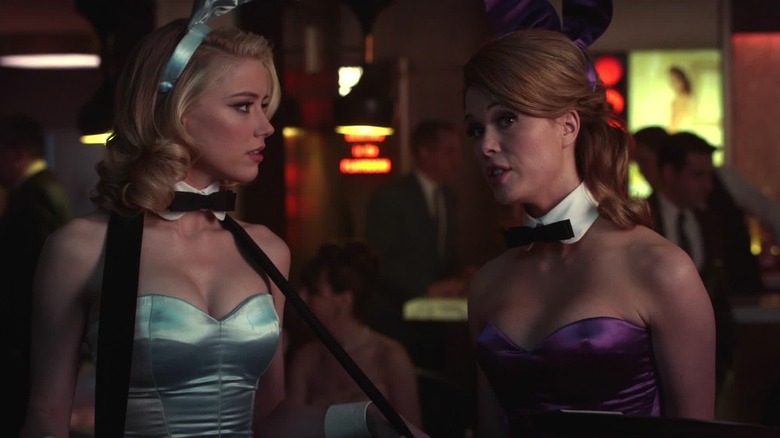 Amber Heard (Maureen), wearing a light blue Playboy bunny costume and carrying a cigarette tray, looks to the right at Alice (Leah Renee), in the middle of speaking in a purple bunny costume, inside a dimly lit club in The Playboy Club