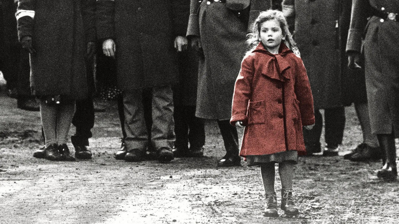 A girl in a red coat (Oliwia Dąbrowska) stands on a gravelly road with a surprised expression, the bodies of men in suits and an SS officer behind her in black and white in Schindler's List