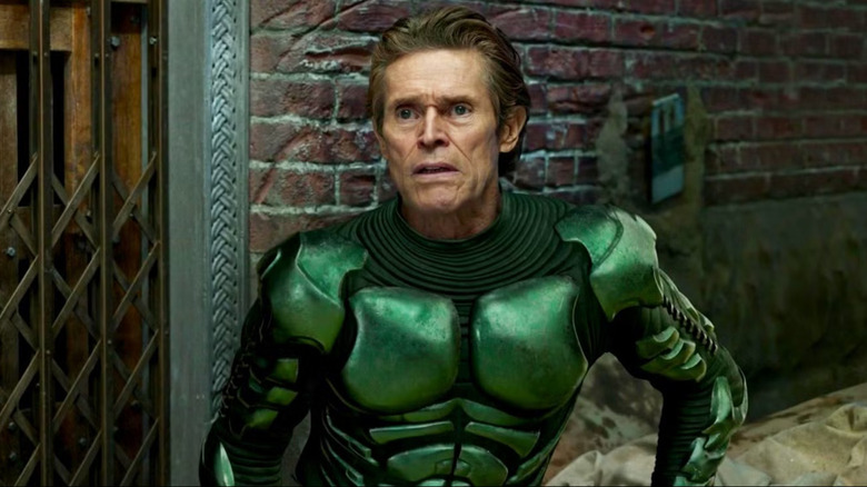 Norman Osborn (Willem Dafoe) leans against a brick wall beside a vintage elevator gate, wearing his Green Goblin suit and a concerned expression in Spider-Man: No Way Home